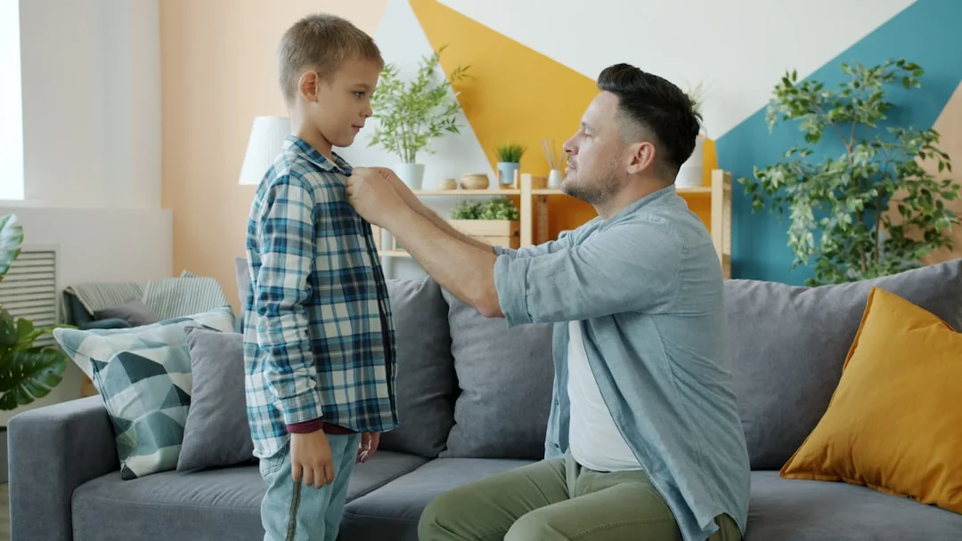 Father buttons son's plaid shirt while sitting on couch. (Fot. Vitaly Gariev / Unsplash)
